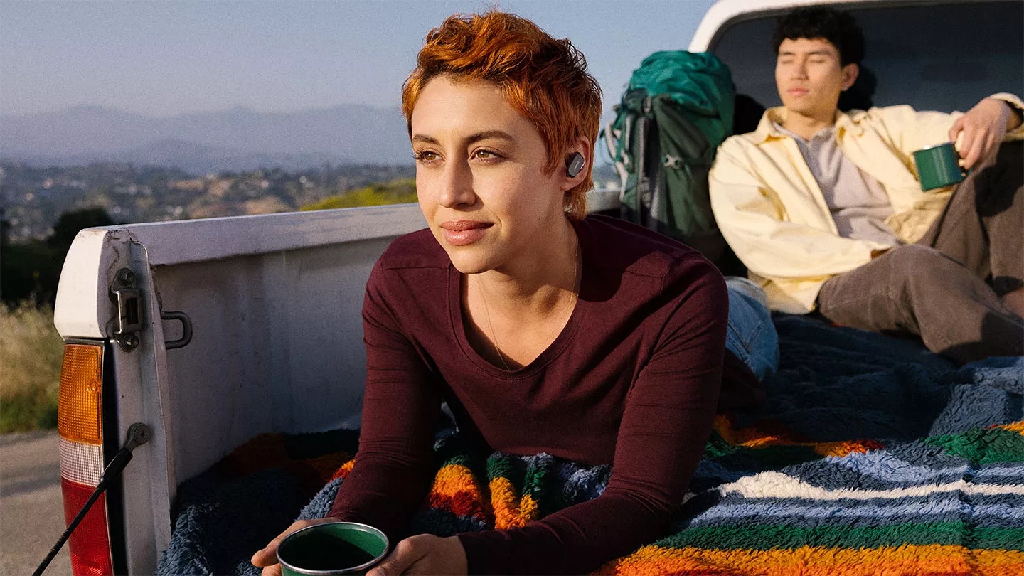 Couple sitting in back of pickup truck drinking coffee and woman listening to QuietComfort Earbuds II