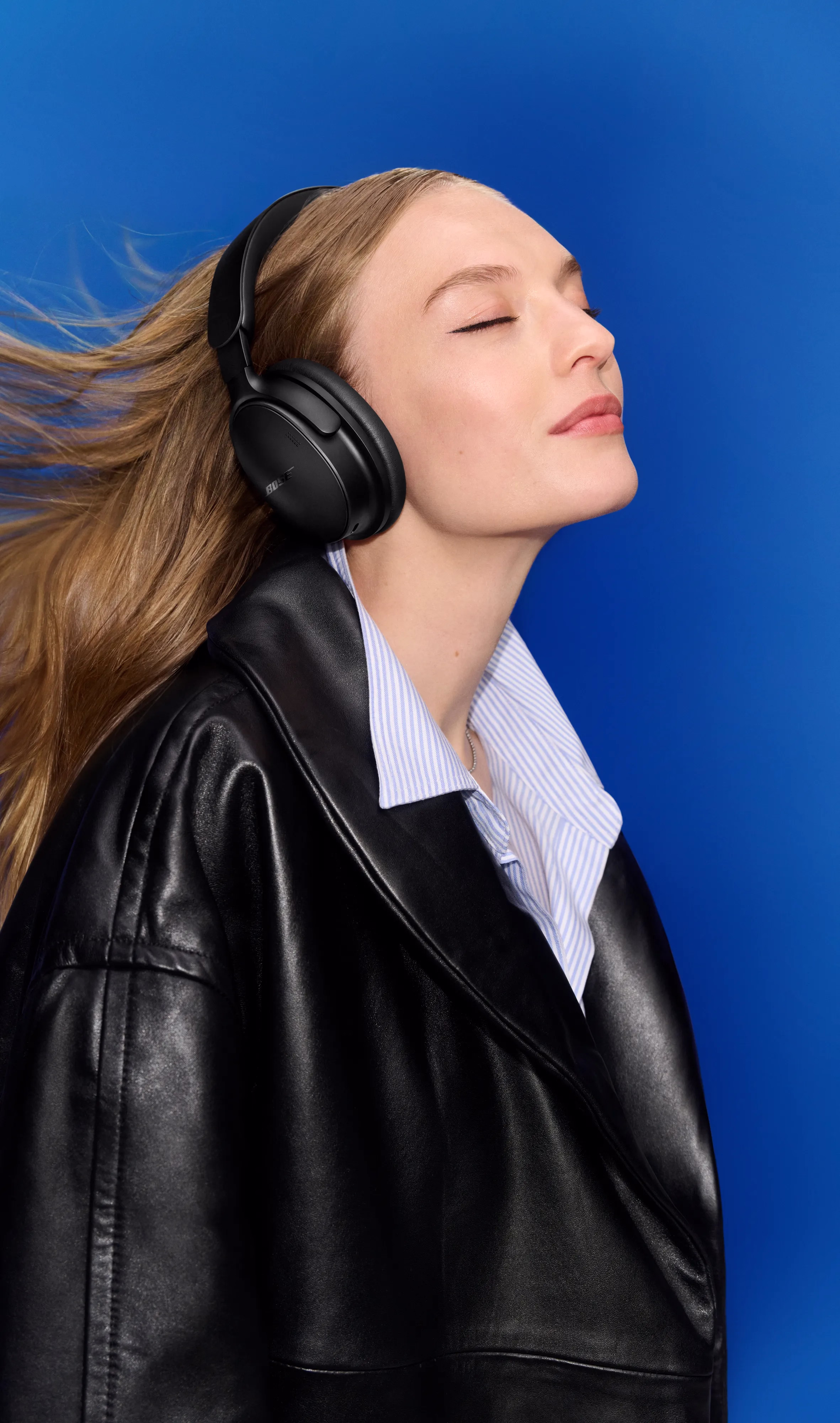 Woman wearing Black QuietComfort Headphones (2nd Gen) with her eyes closed against a vibrant blue background, hair flowing to the side.