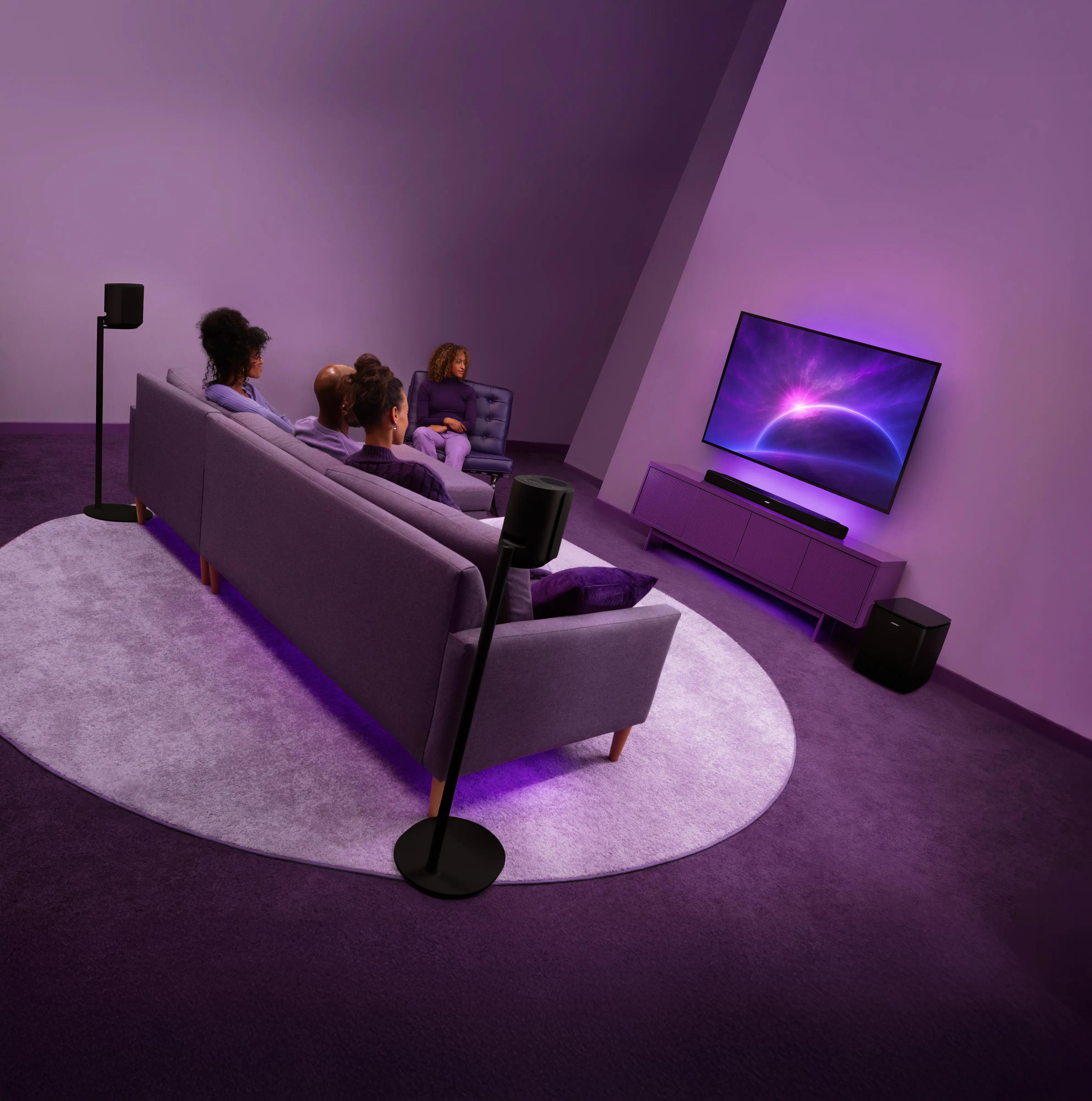 Modern home theater setup featuring the Lifestyle Ultra Soundbar below a wall-mounted TV, Lifestyle Ultra Speakers mounted on Lifestyle Ultra Speaker Floor Stands surrounding the seating area, and a Lifestyle Ultra Subwoofer beside the media console.