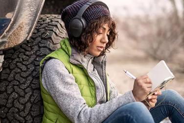 Woman leaning against a truck wearing Bose QuietComfort 45 headphones writing in a journal