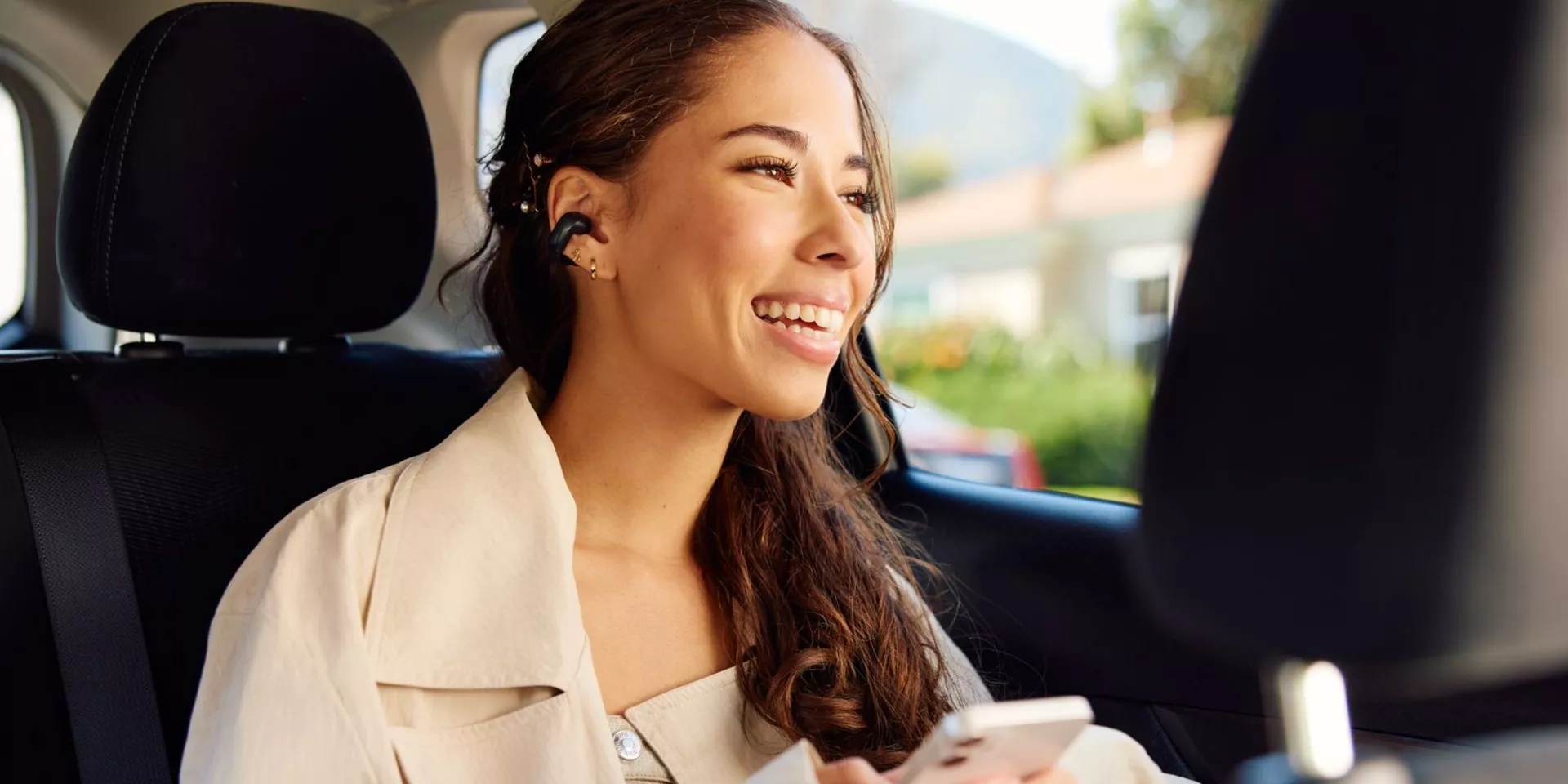 Woman wearing Bose Ultra Open Earbuds while sitting in the back of a car listening to music from her phone