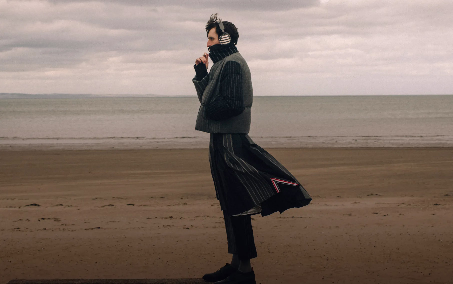 Man wearing Bose &times; Thom Browne QuietComfort Ultra Headphones (2nd Gen) while standing on a beach. 