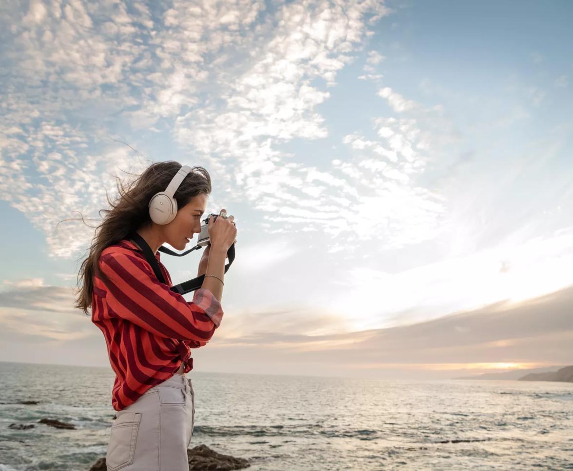 A woman photographing the sunrise at a beach wearing Bose QuietComfort 45 headphones