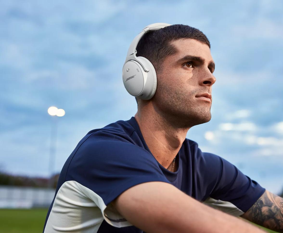 Christian Pulisic sitting on a soccer field wearing QuietComfort 45 headphones