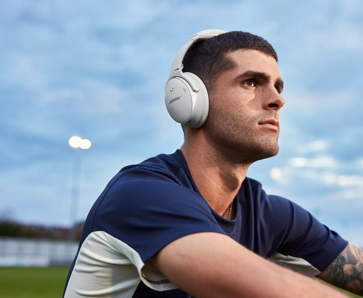 Christian Pulisic sitting on a soccer field wearing QuietComfort 45 headphones