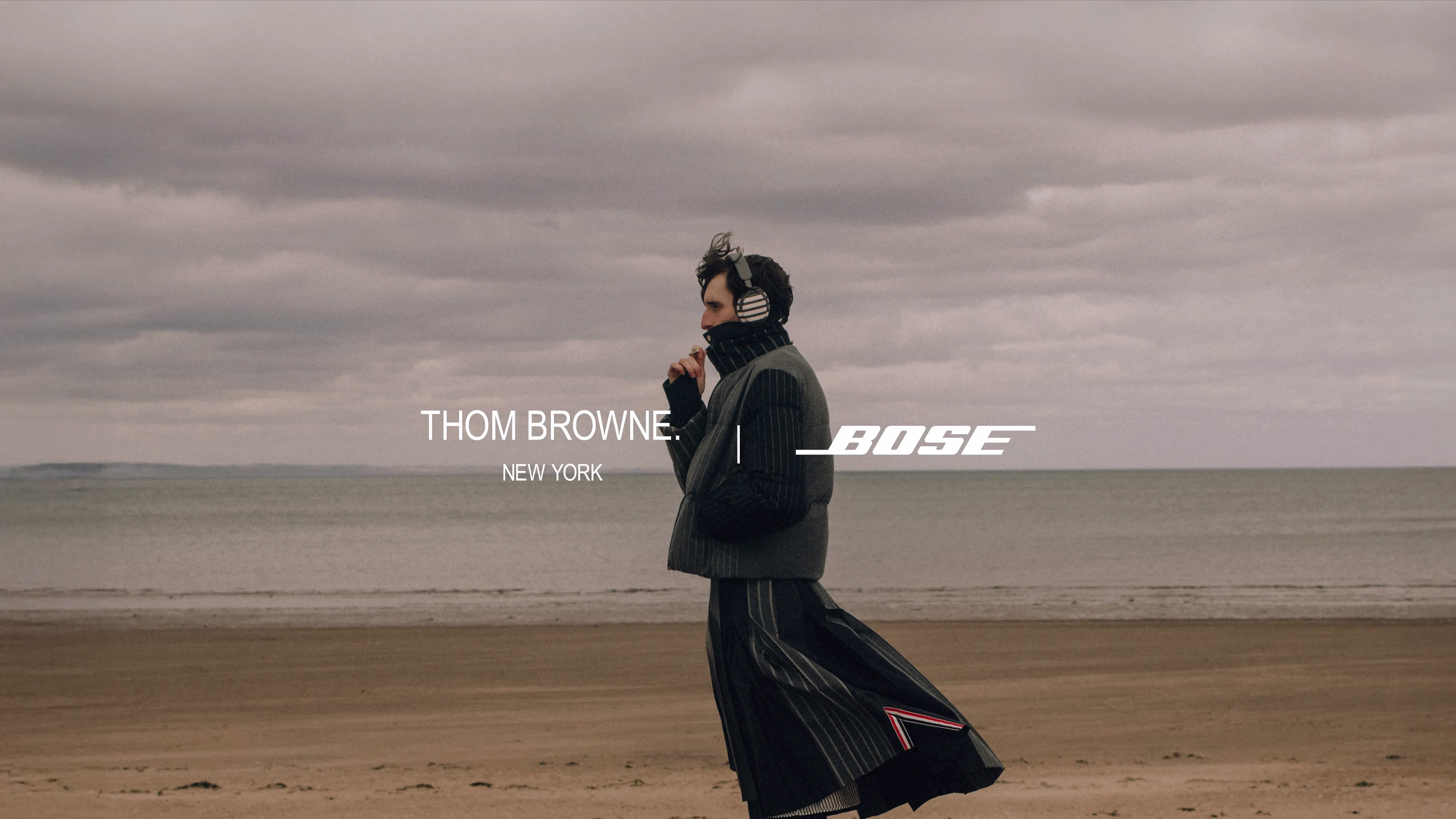 Logo Bose et Thom Browne, et homme portant le casque QuietComfort Ultra (2e&nbsp;g&eacute;n&eacute;ration) Bose &times; Thom Browne (2e&nbsp;g&eacute;n&eacute;ration), debout sur une plage.