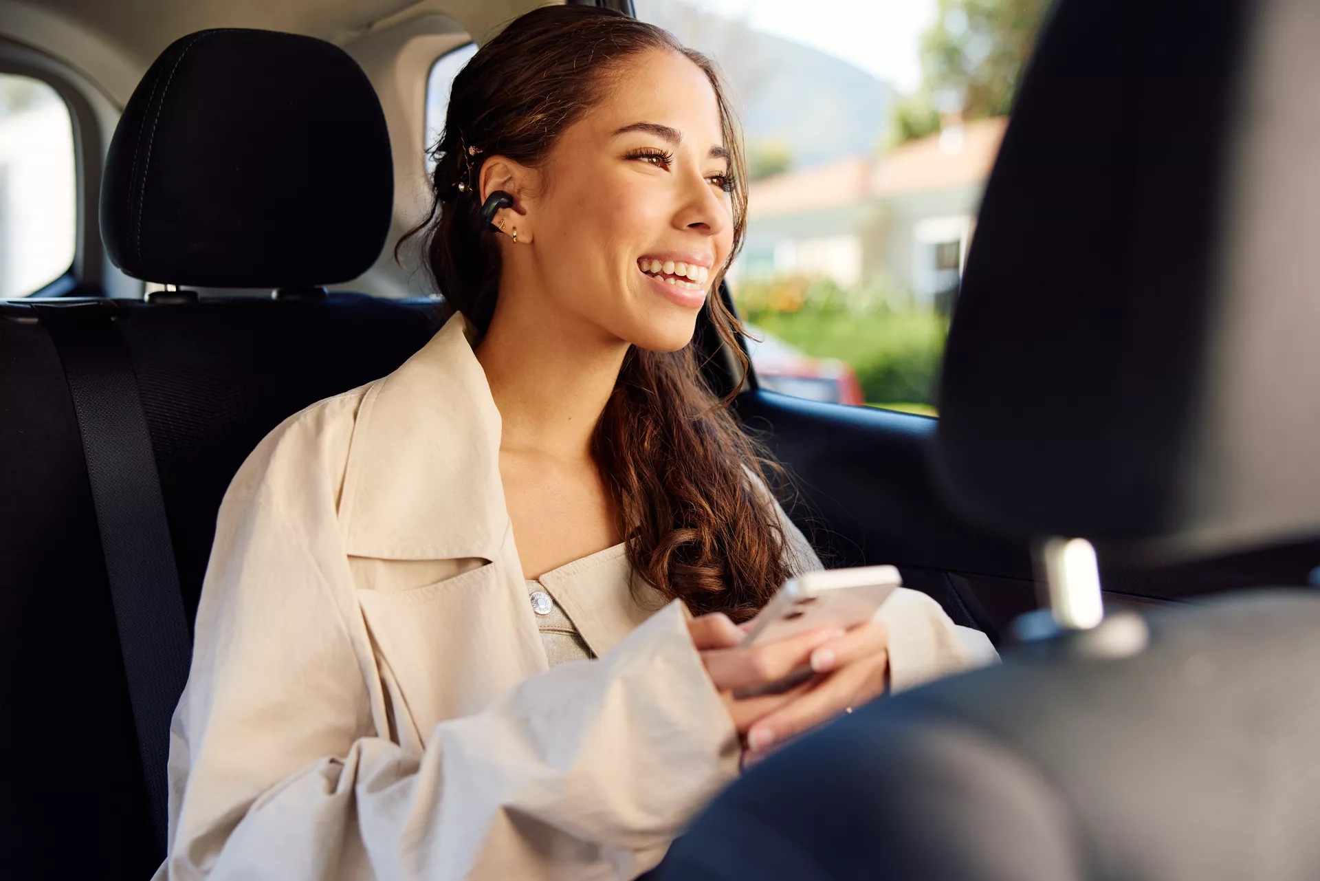 Woman wearing Bose Ultra Open Earbuds while sitting in the back of a car listening to music from her phone