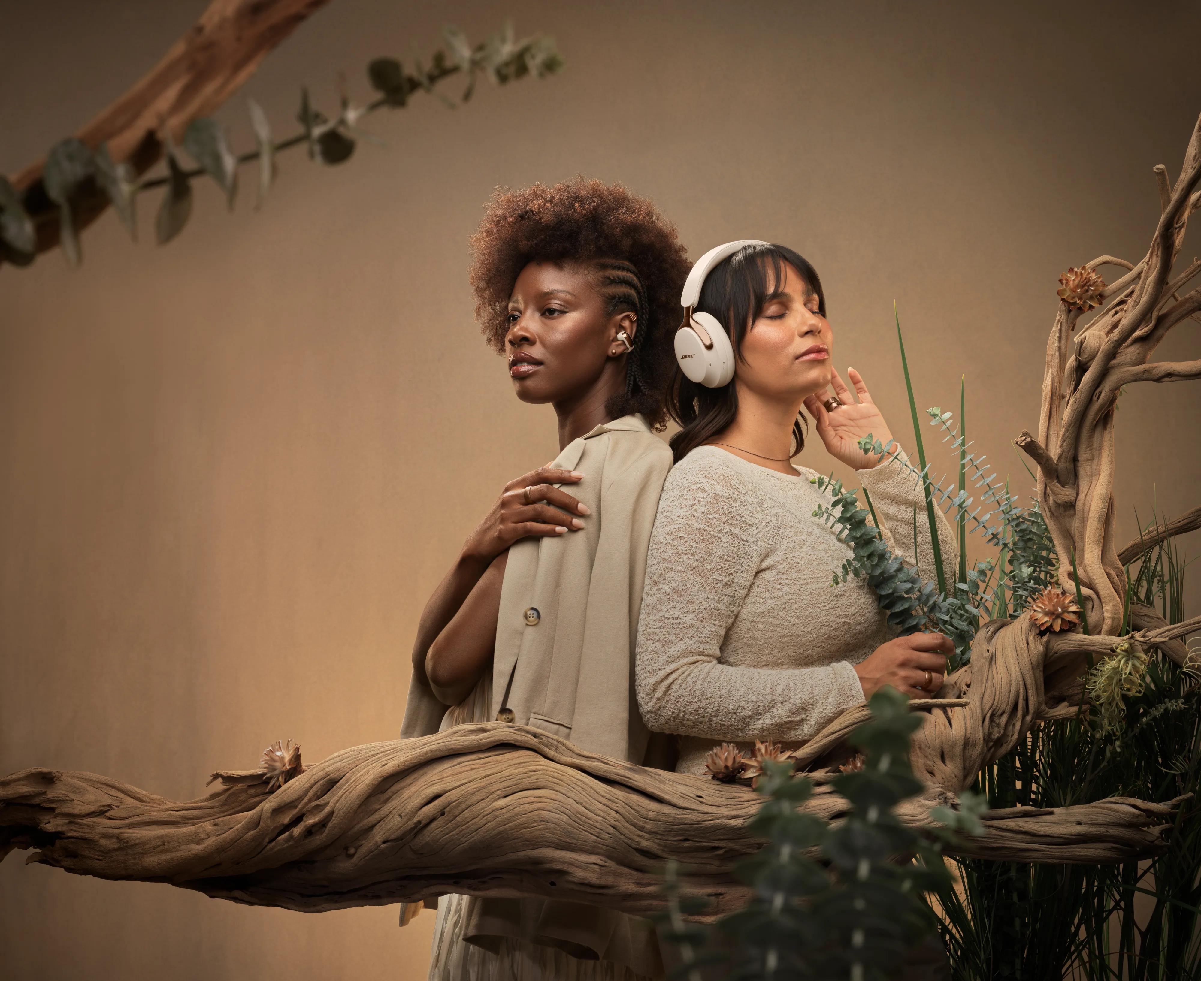 Two women wearing QuietComfort Ultra Headphones LE (2nd Gen), and Ultra Open Earbuds LE in Driftwood Sand, posed back-to-back in a warm, natural setting with organic textures, highlighting the cohesive neutral-toned design.