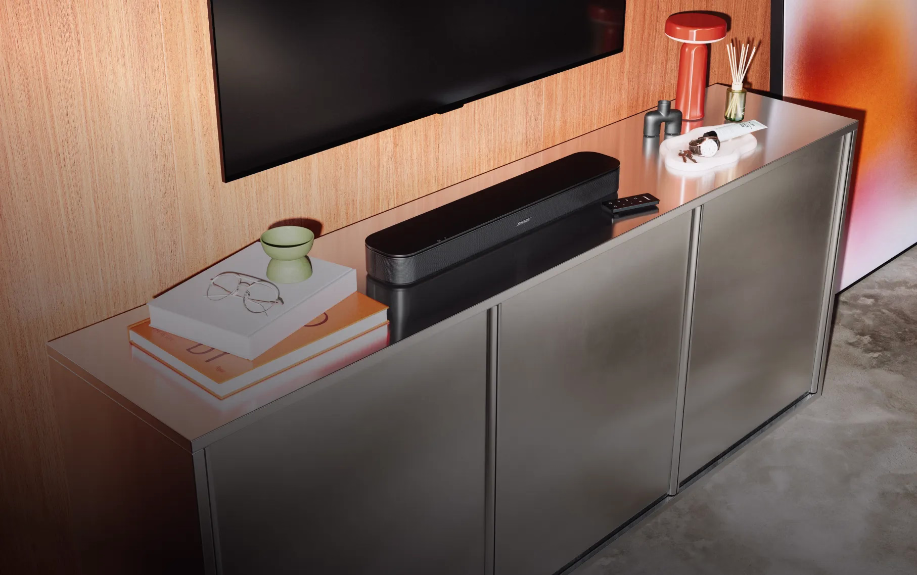 Bose TV Speaker (2nd Gen) and its remote on a TV console, with a TV mounted on the wall above them.