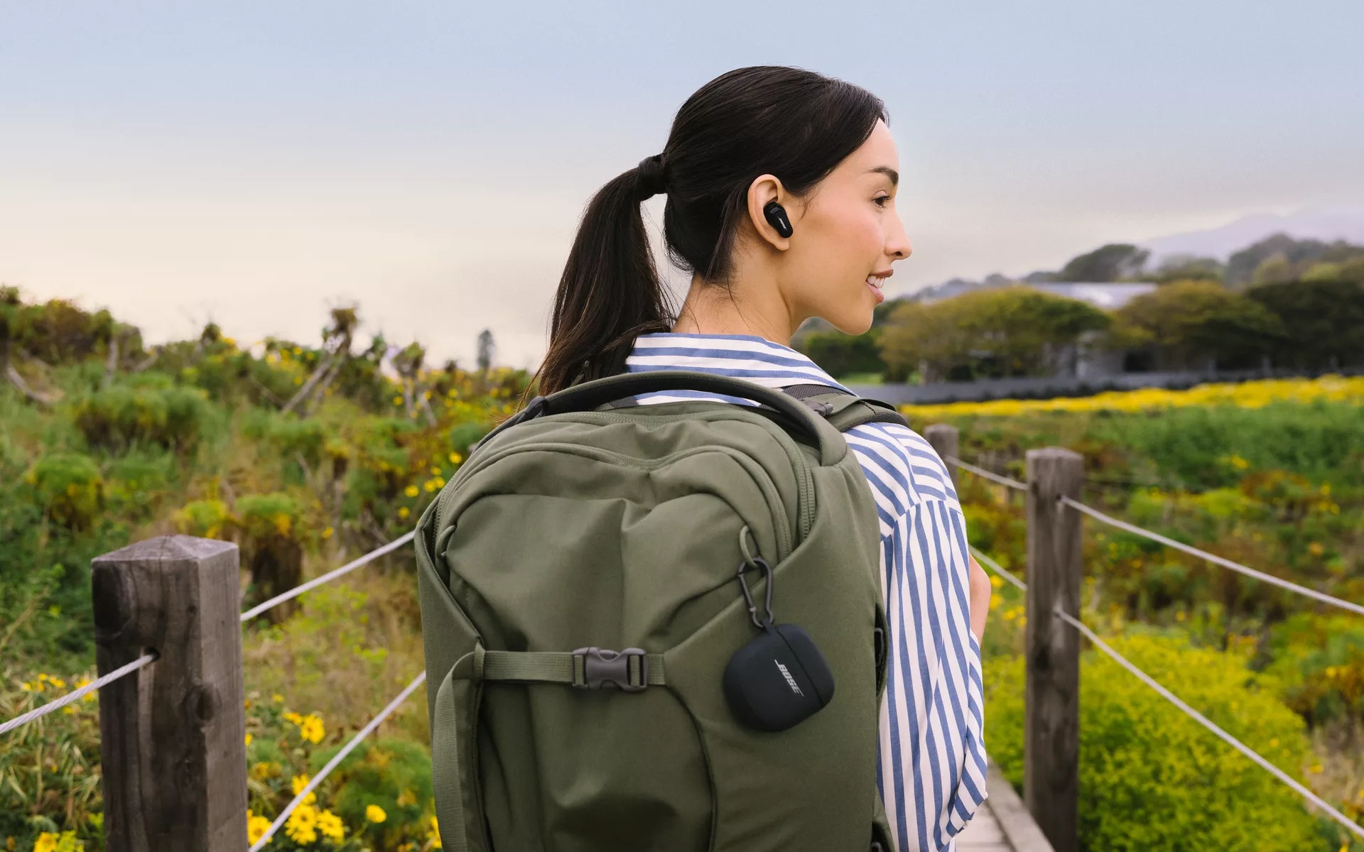 Woman wearing QuietComfort Earbuds II while hiking, with the charging case inside the QuietComfort Silicone Case Cover attached to her backpack