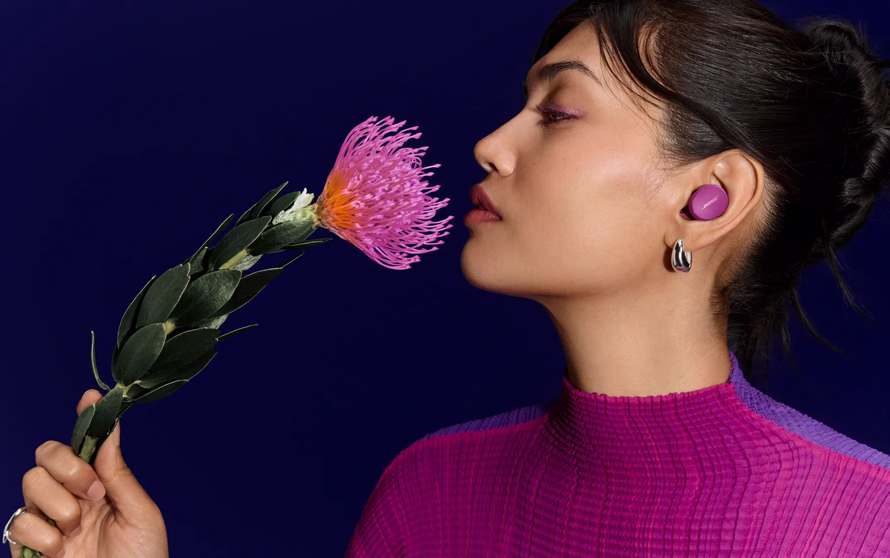 Woman listening to music from Bose QuietComfort Earbuds in Rosewood Mauve while wearing a bright pink sweater smelling a pink Pincushion flower.