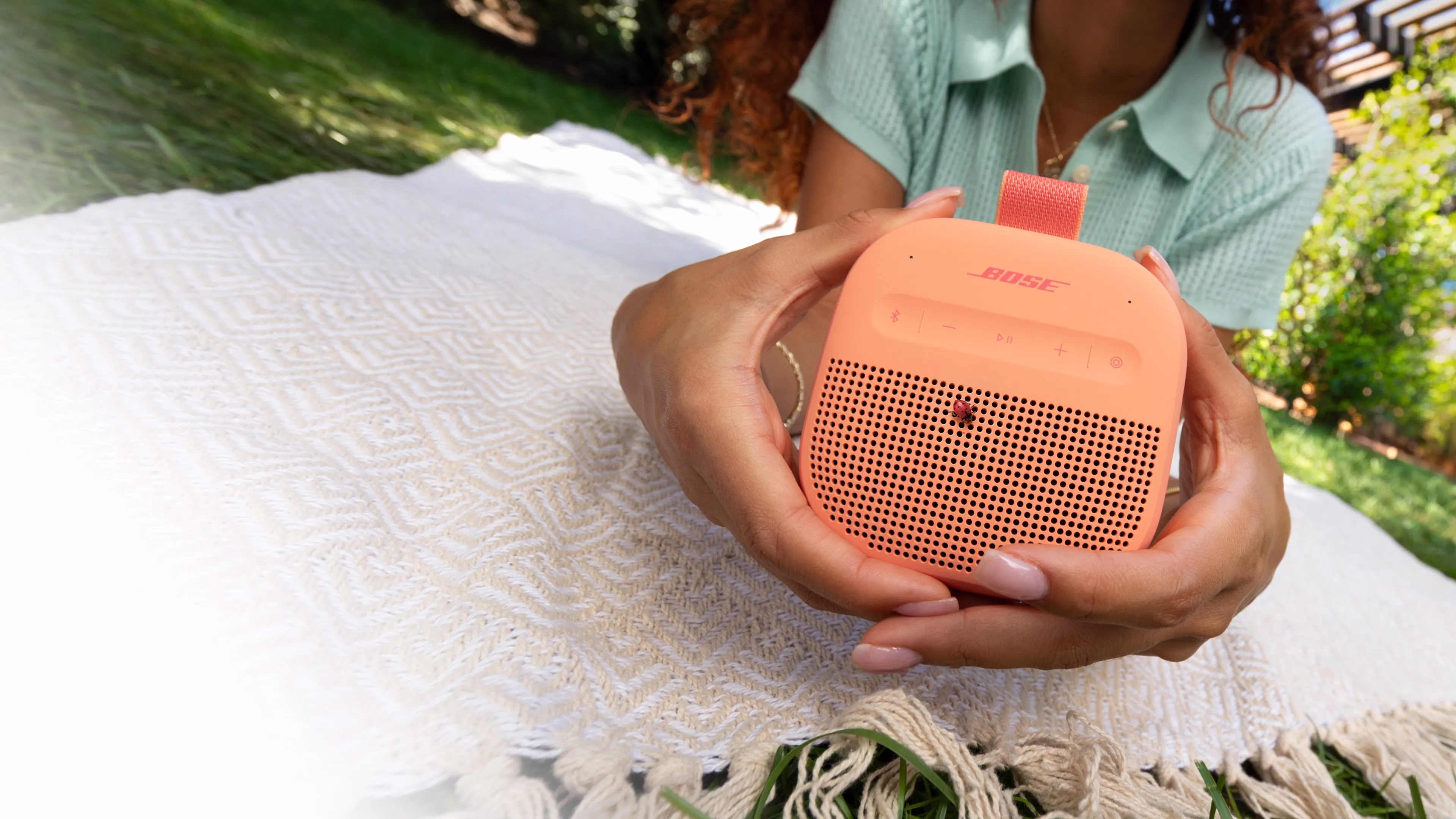 Woman holding a Bose SoundLink Micro Portable Speaker (2nd Gen) while lying on a blanket on the grass.