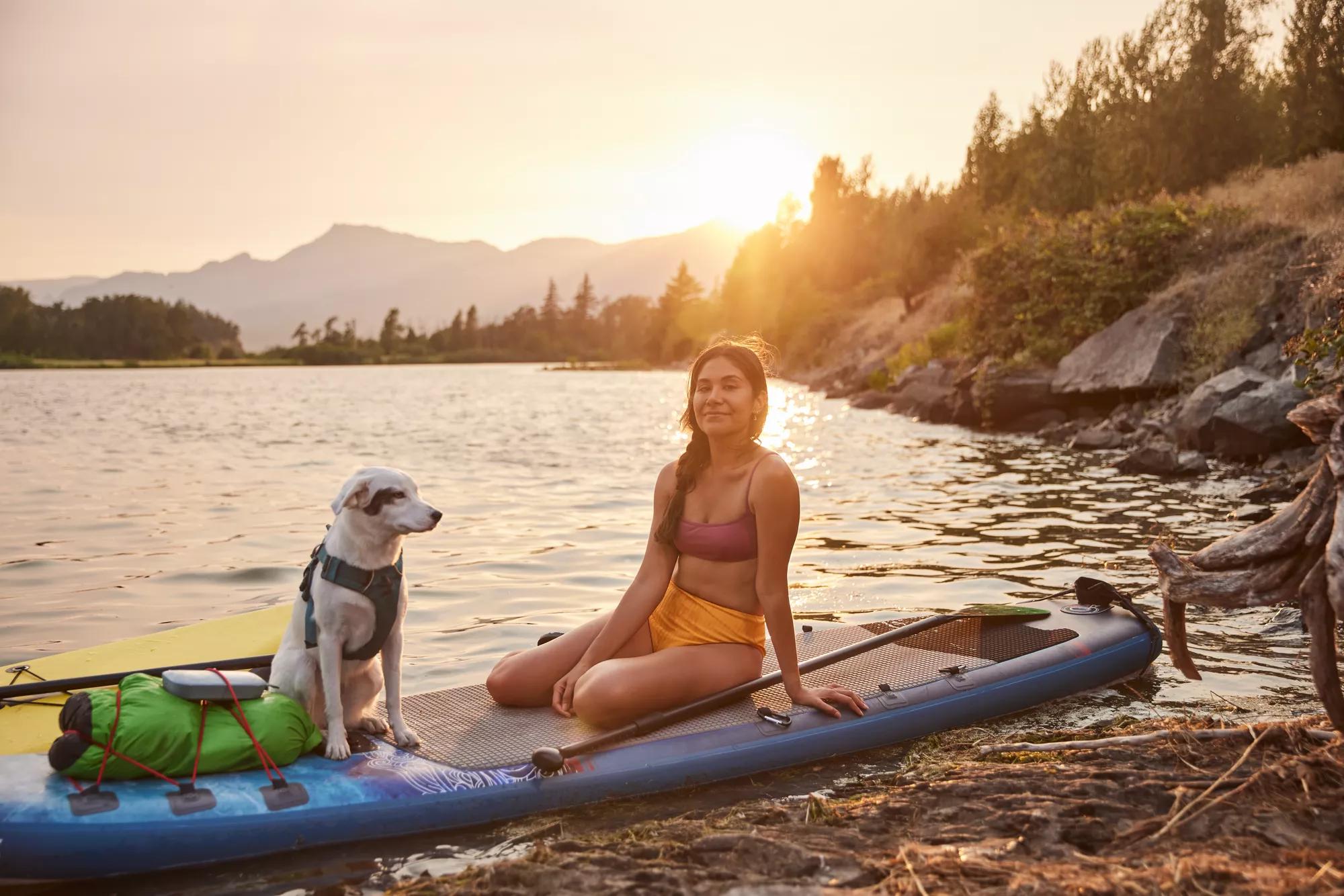 A woman and her dog on a paddleboard by the lakeshore listening to music on a Bose SoundLink Flex portable Bluetooth speaker