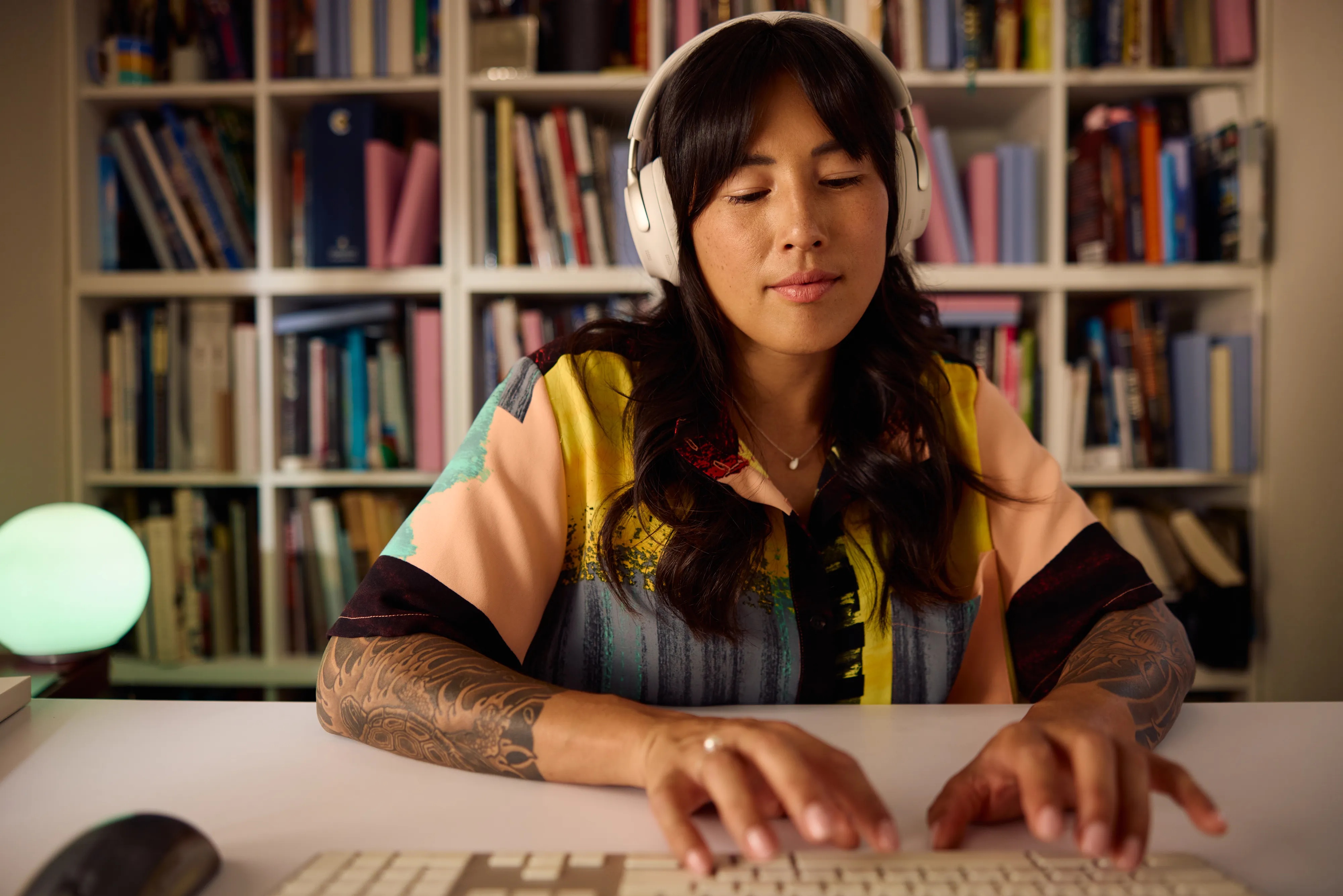 A woman wearing white smoke QuietComfort Ultra Headphones while typing on a keyboard at a desk, with a library in the background.