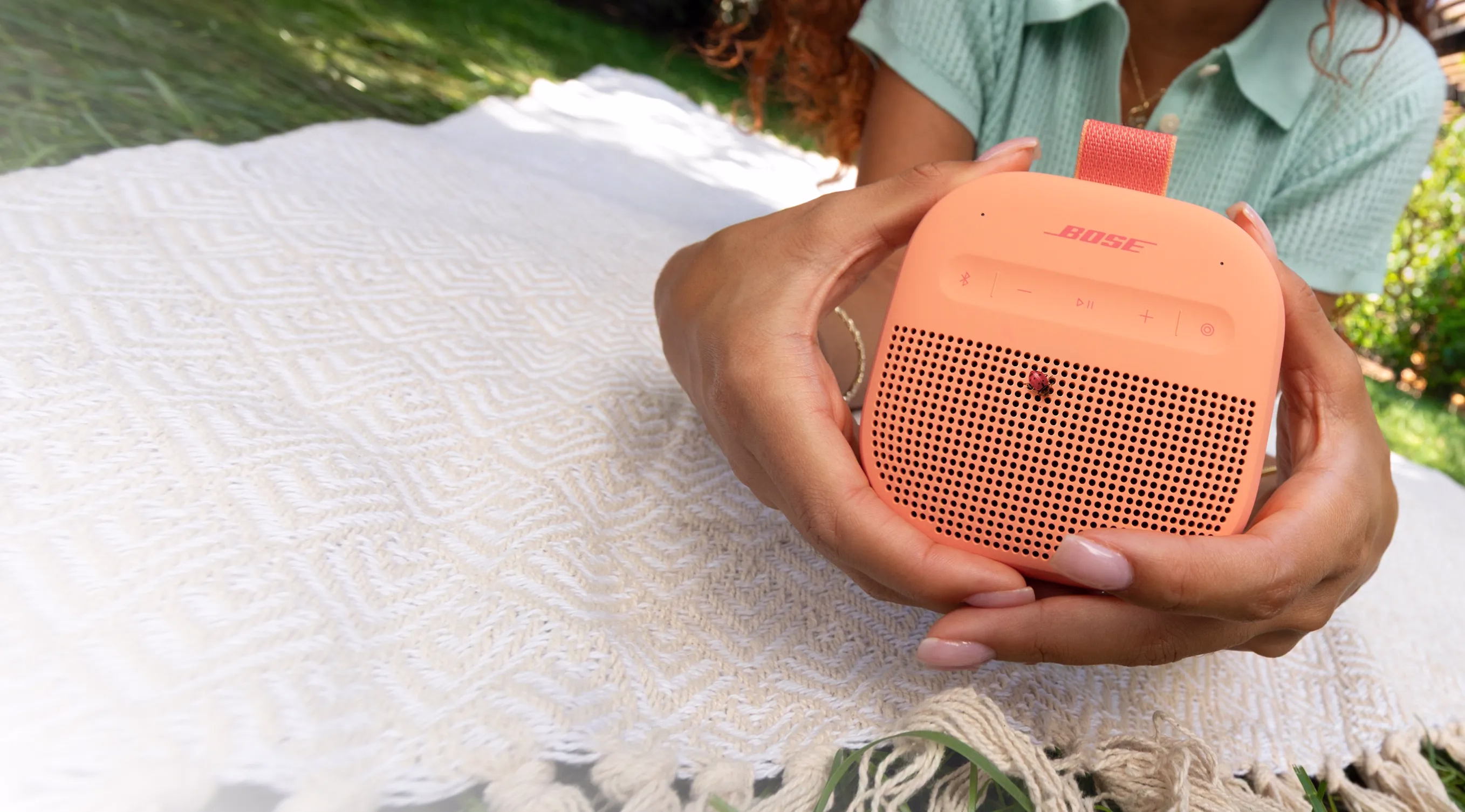 Woman lying down on a blanket on the grass holding a Bose SoundLink Micro Portable Speaker (2nd Gen) in Sunset Peach.