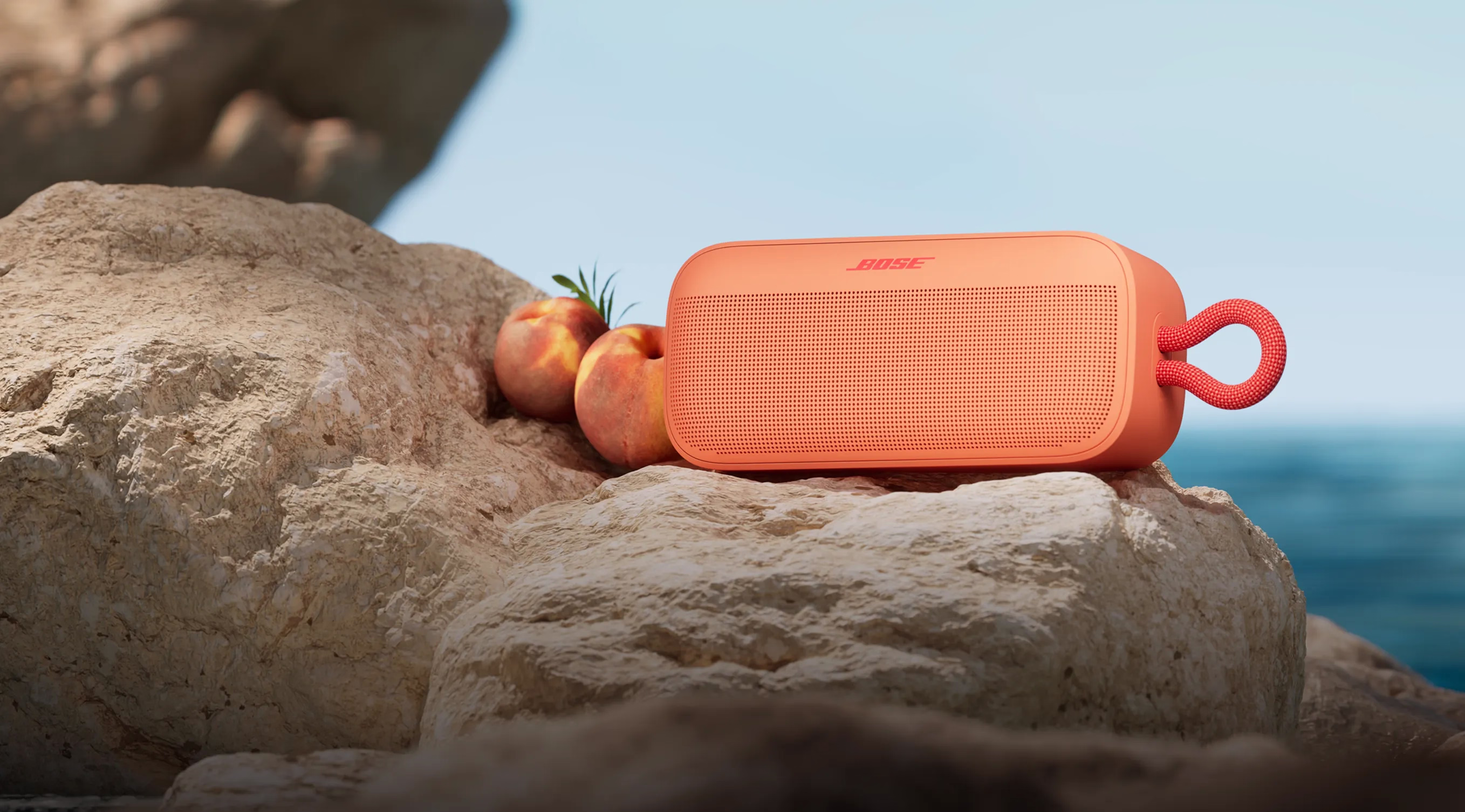 ビーチで、サンセットピーチ色のSoundLink Plusポータブルスピーカーに寄りかかる桃。