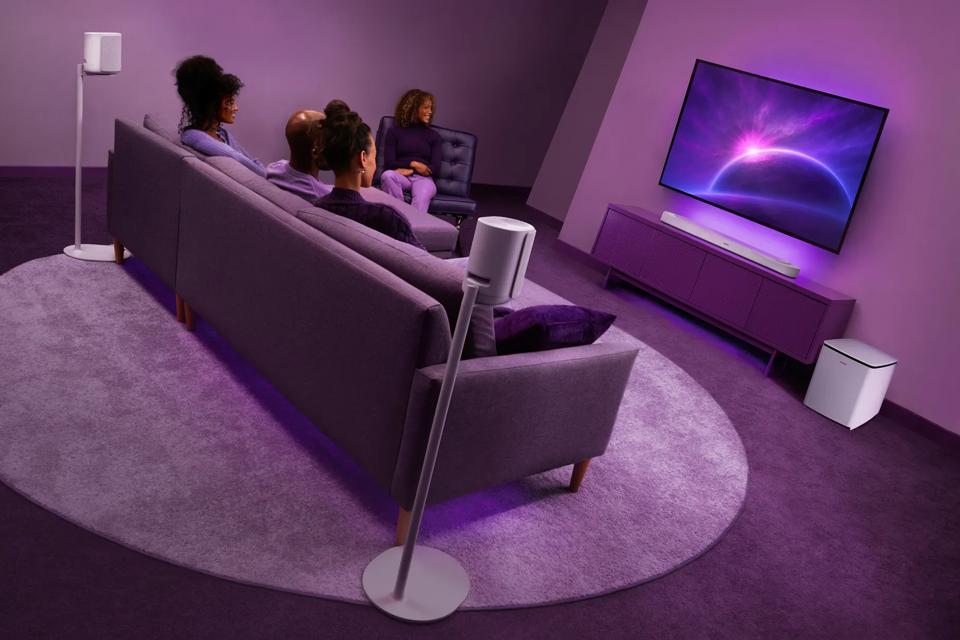 Friends sitting in a living room watching TV with a Bose Lifestyle Ultra Soundbar, Lifestyle Ultra Speakers on floor stands, and Lifestyle Ultra Subwoofer.