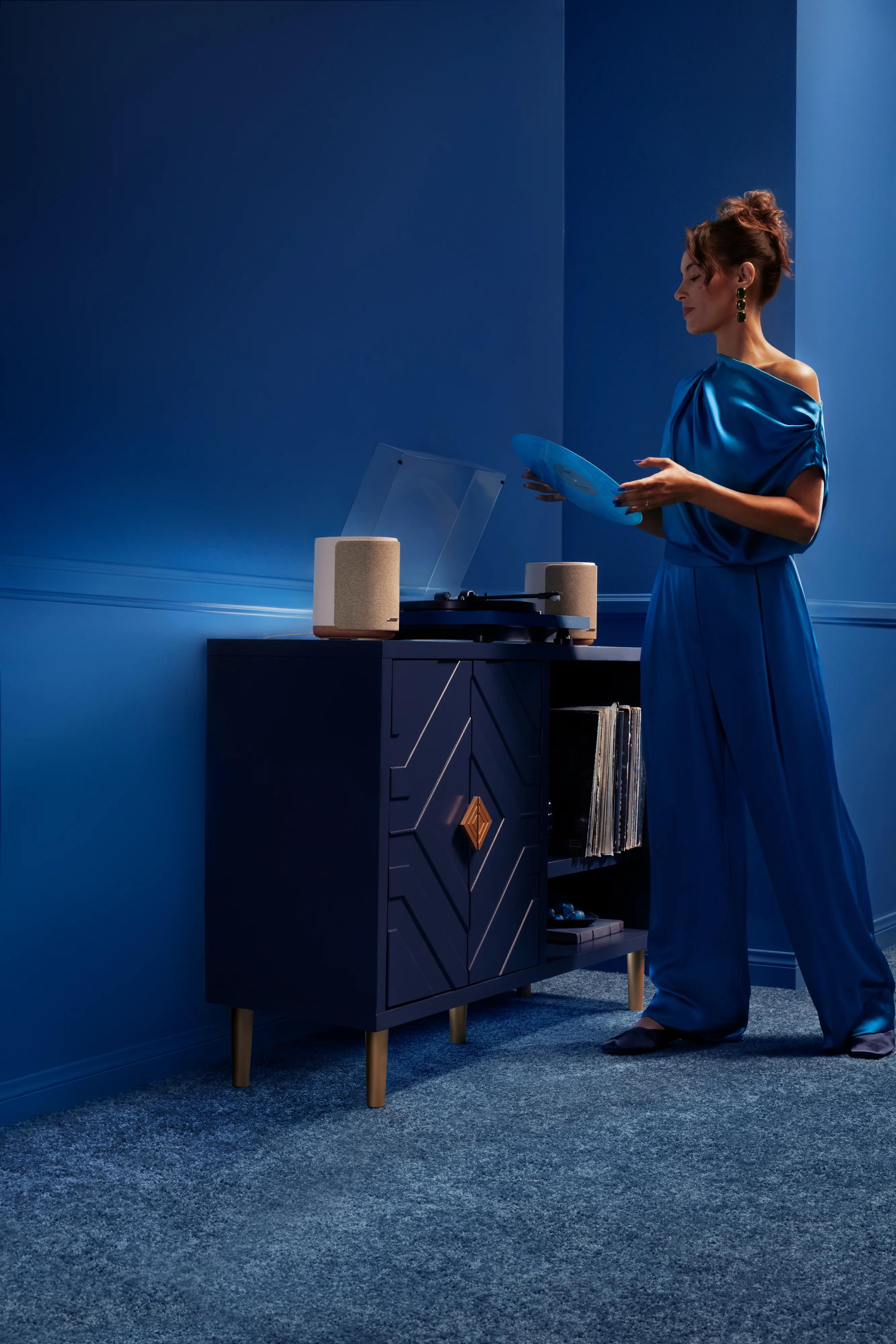 Woman holding a vinyl record beside Lifestyle Ultra Speakers and a record player on a modern media cabinet in a softly lit room.