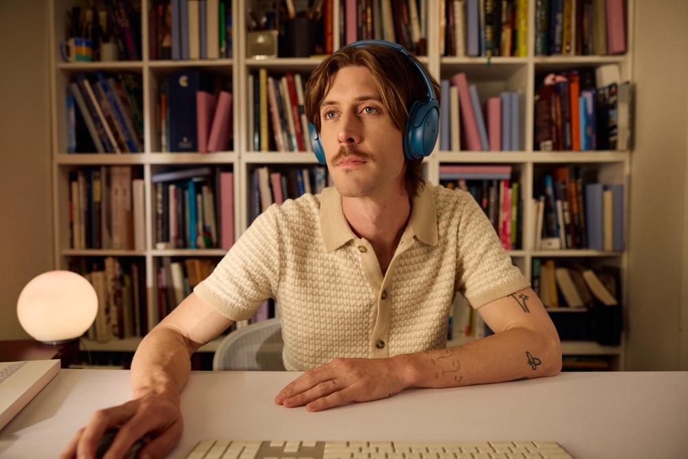 A man wearing Twilight Blue QuietComfort Ultra Headphones LE while typing on a keyboard at a desk, with a library in the background.