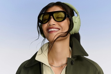 Woman wearing Eucalyptus Green Bose QuietComfort Headphones (2nd Gen) while smiling with a light blue background.