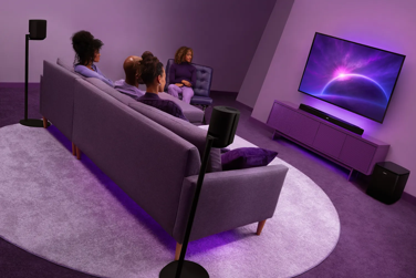 Modern home theater setup featuring the Lifestyle Ultra Soundbar below a wall-mounted TV, Lifestyle Ultra Speakers mounted on Lifestyle Ultra Speaker Floor Stands surrounding the seating area, and a Lifestyle Ultra Subwoofer beside the media console.