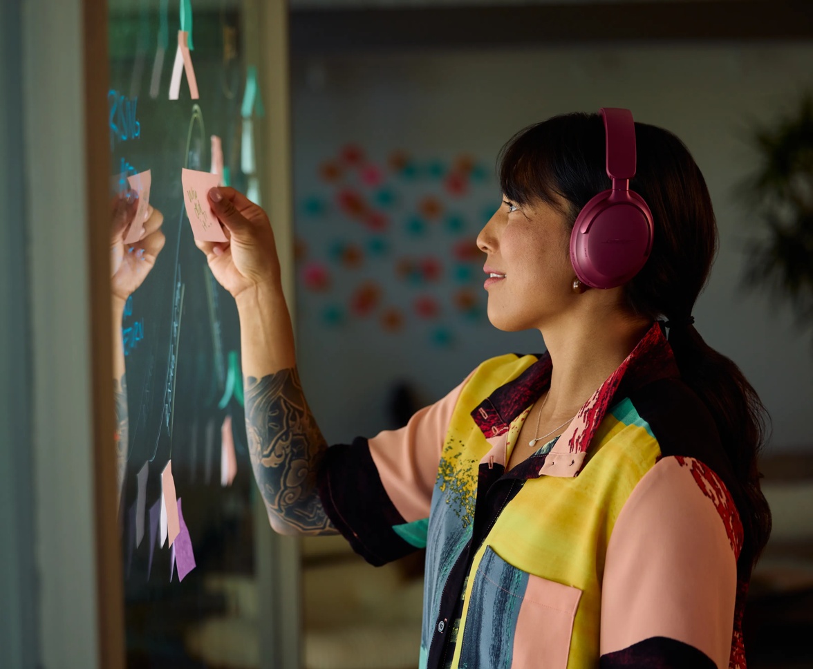 A woman wearing Deep Plum QuietComfort Ultra Headphones LE while writing in a vinyl board 