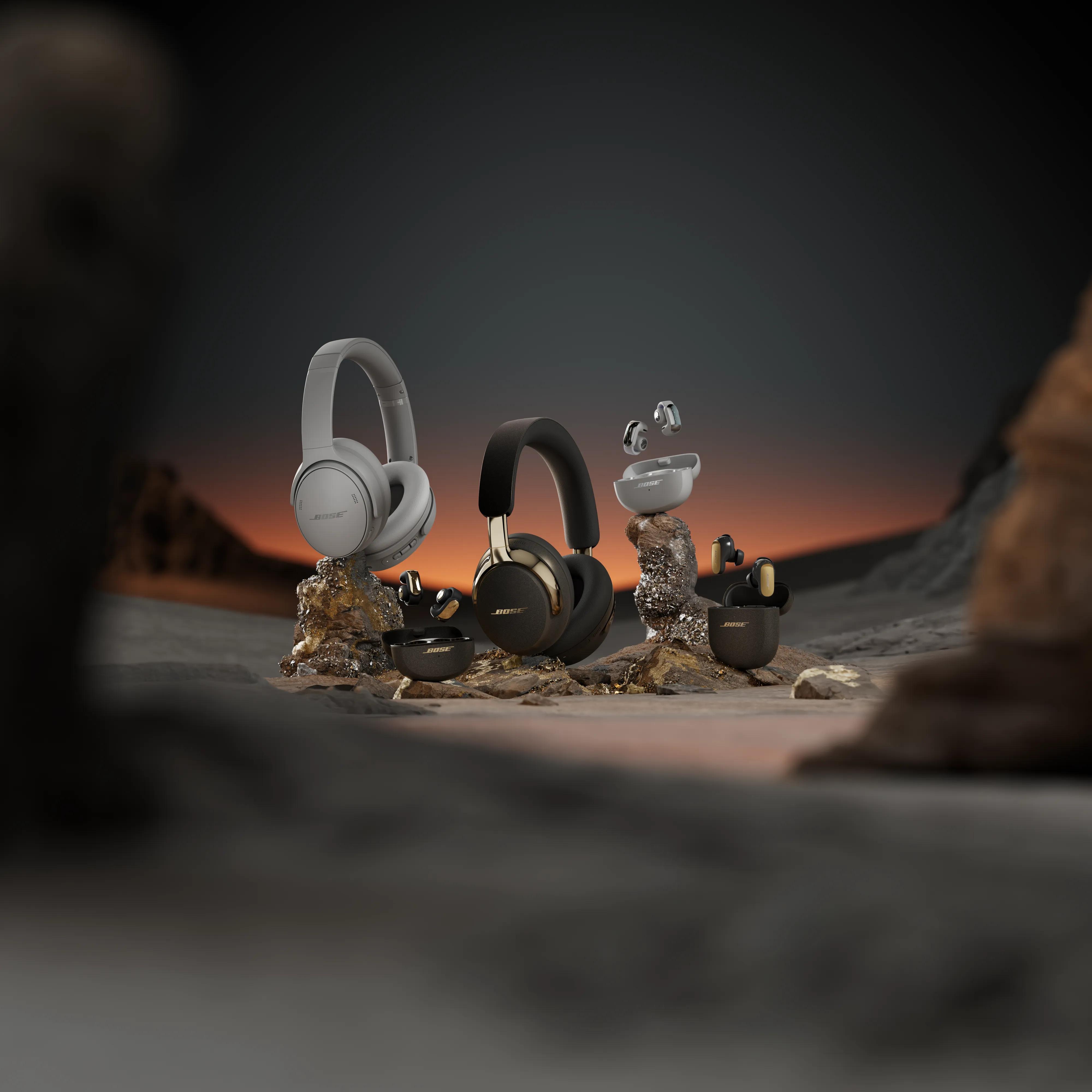 QuietComfort Headphones LE, QuietComfort Ultra Earbuds LE (2nd Gen), QuietComfort Ultra Headphones LE (2nd Gen), and Ultra Open Earbuds LE displayed on rocky surfaces in a desert-like setting at sunset.