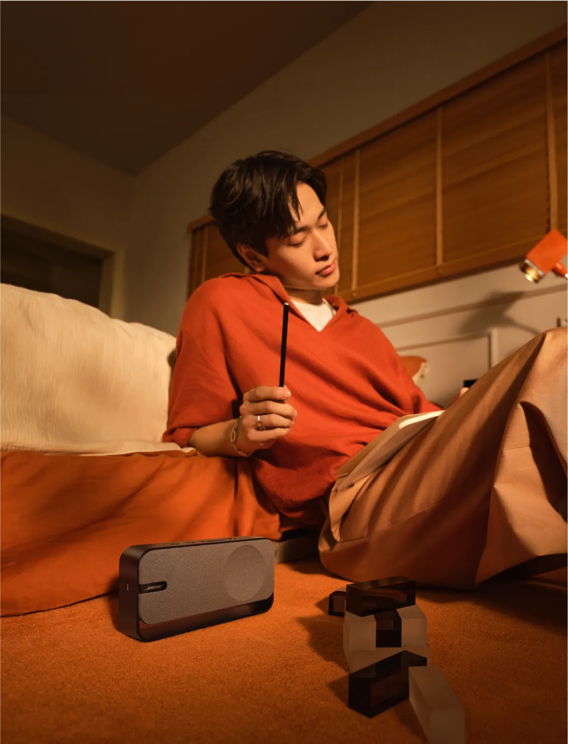Jeune homme assis sur le sol dans sa chambre, appuy&eacute; contre son lit &eacute;tudiant, &eacute;coutant un haut-parleur Bose SoundLink Home Bluetooth.