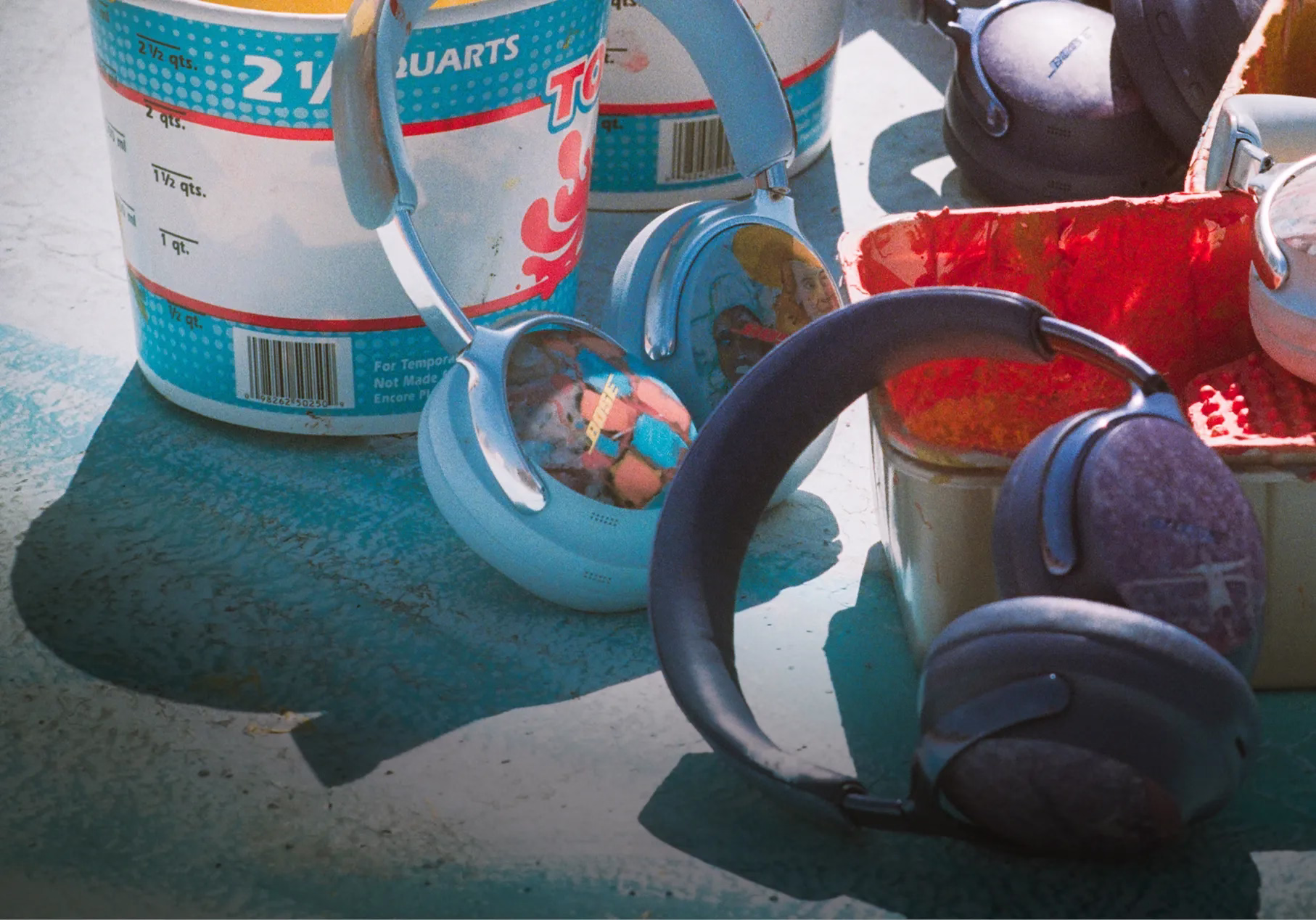 Bose &times; KidSuper QuietComfort Ultra Headphones (2nd Gen) in Moonstone Blue and Midnight Violet on a rooftop with paint cans and paint trays.