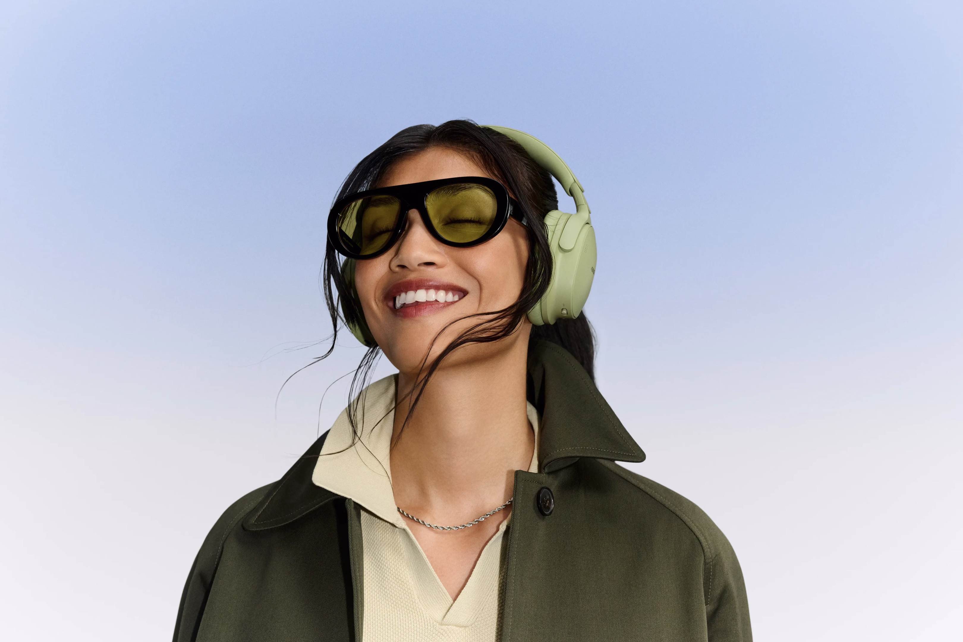 Woman wearing Eucalyptus Green Bose QuietComfort Headphones (2nd Gen) while smiling with a light blue background.
