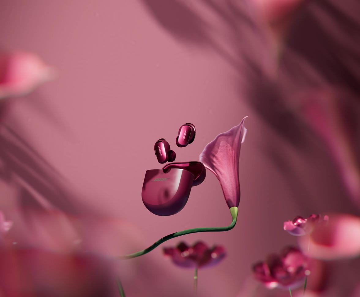 Bose QuietComfort Ultra floating above an open charging case beside a pink calla lily on a matching background.