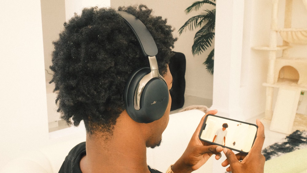 A man wearing black Bose QuietComfort Ultra Headphones (2nd Gen) while listening to a video on his smartphone. 