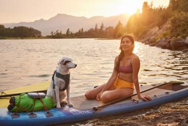 Ana Alarcón and her dog sitting on her paddleboard by a lake with a SoundLink Flex Bluetooth speaker attached to her backpack