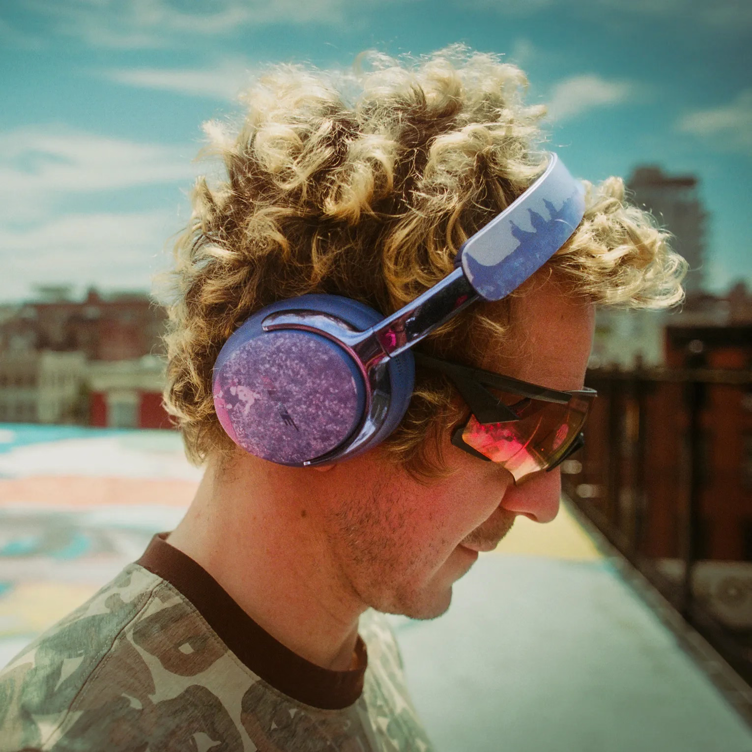 Side profile of KidSuper wearing Midnight Violet Bose &times; KidSuper QuietComfort Ultra Headphones (2nd Gen) on the hand-painted rooftop of a city building.