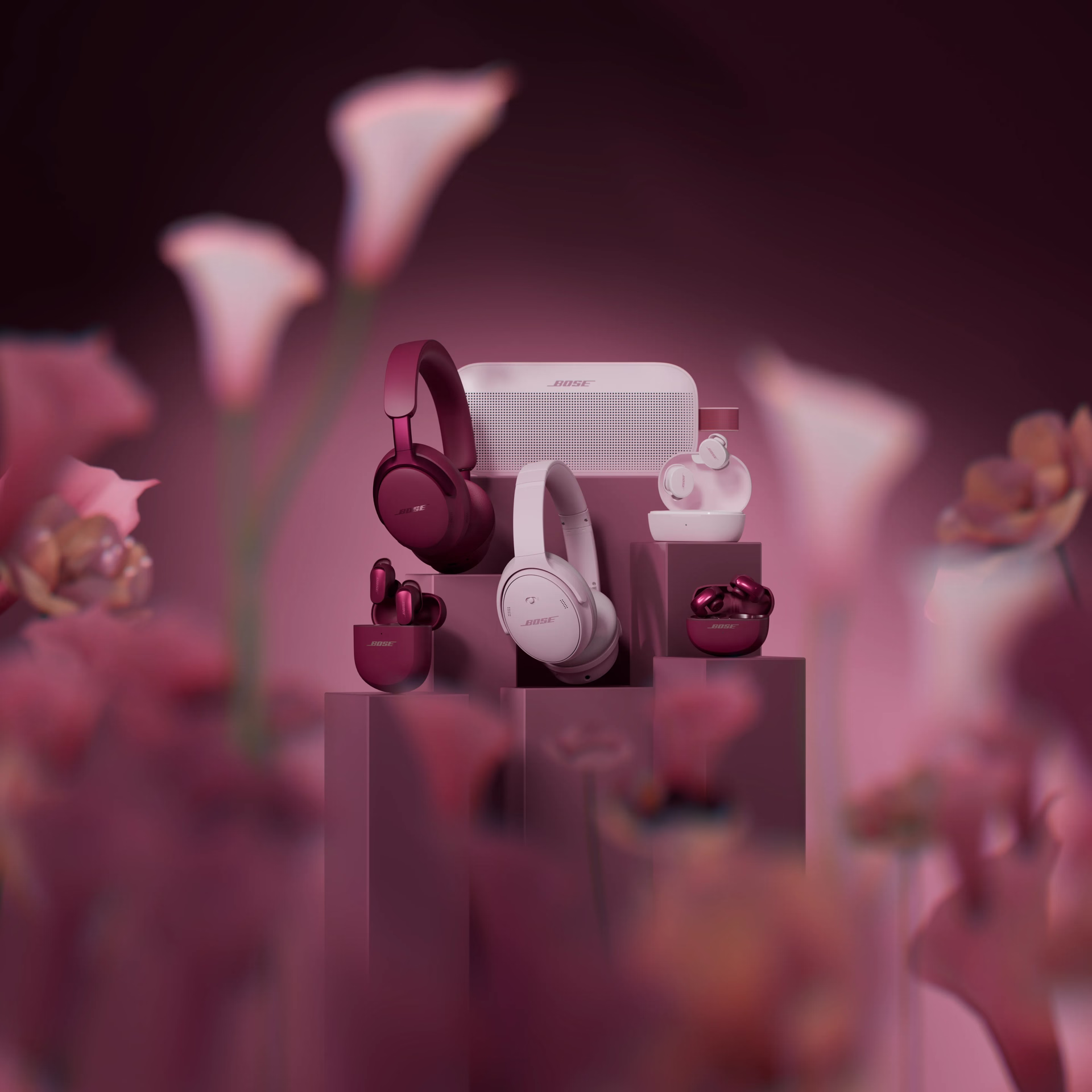 QuietComfort Headphones LE, QuietComfort Ultra Headphones LE, QuietComfort Ultra Earbuds (2nd Gen), SoundLink Flex (2nd Gen) LE, and Ultra Open Earbuds LE displayed on tiered blocks in a clean monochromatic pink setting. 