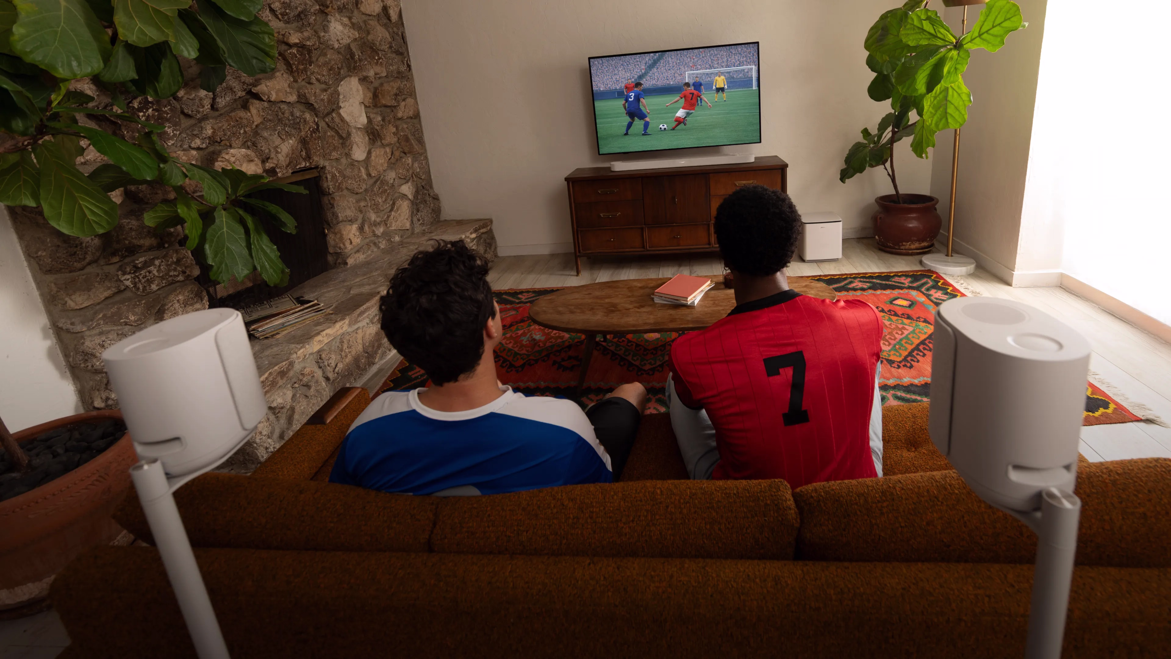 Two friends sitting on a couch in a living room, watching a soccer match on TV with a Bose Lifestyle Ultra Soundbar, Lifestyle Ultra Speakers, and Lifestyle Ultra Subwoofer.